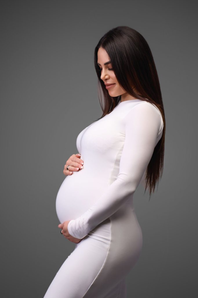 Gravid kvinde i hvid tætsiddende bodysuit med langt sort hår i profil holder om maven mod en grå studiobaggrund