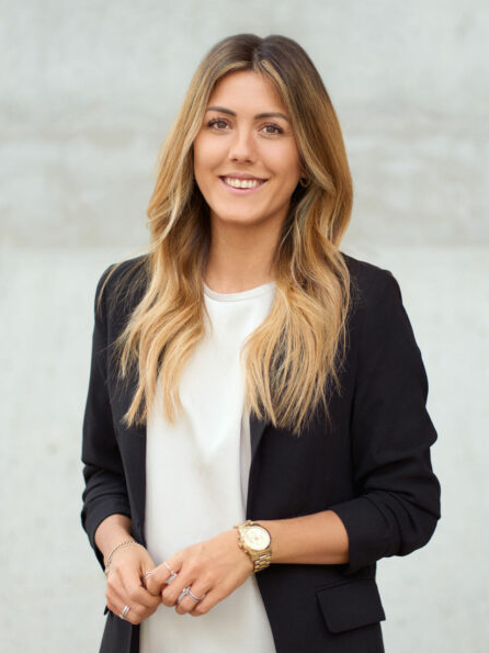 Rundt beskåret medarbejderfoto af kvinde med blondt ombre hår i sort blazer og hvid top, smiler, betonvæg bagved
