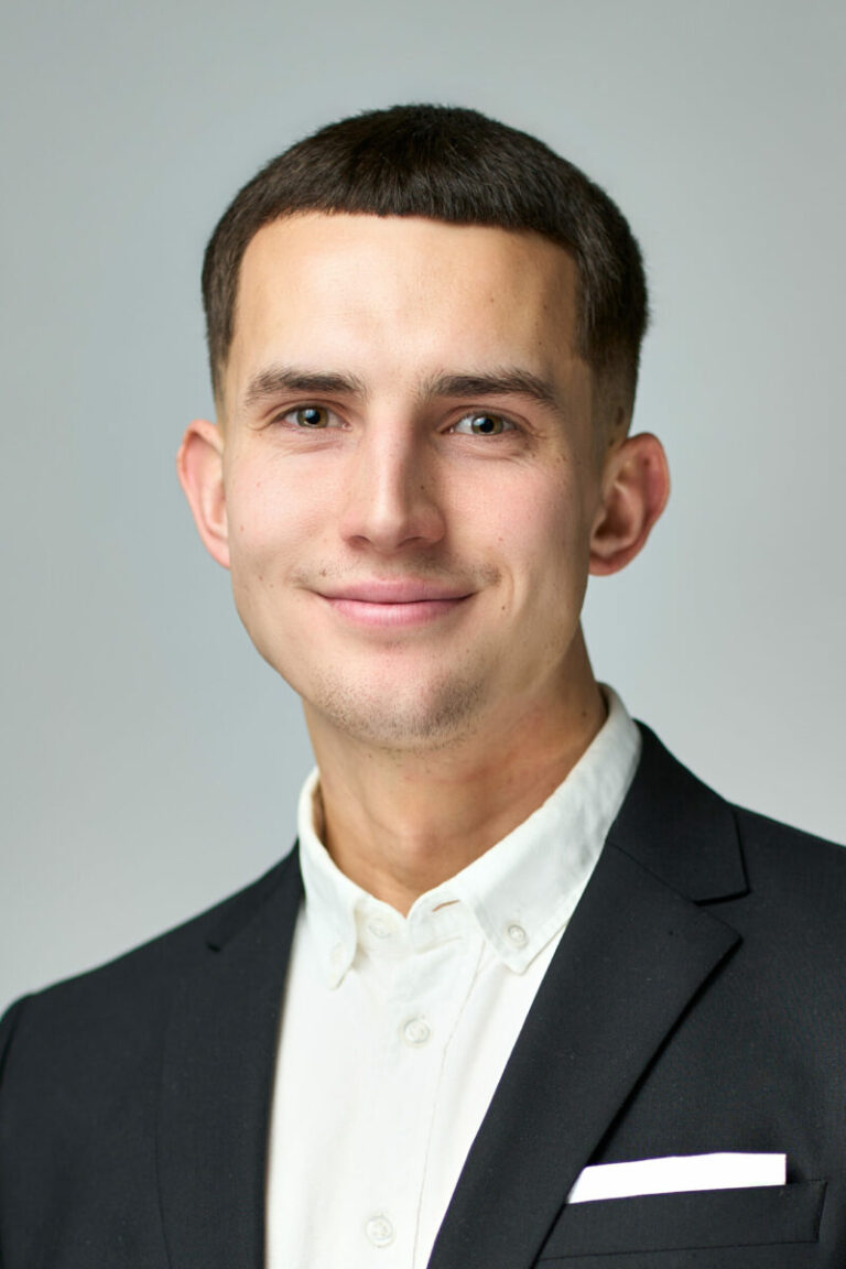 En ung mand med kort mørkt hår, iført sort habitjakke og hvid skjorte uden slips, smiler til kameraet på en lysegrå baggrund, perfekt til et professionelt Linkedin-portræt.