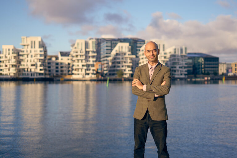 En mand i blazer står med armene over kors ved en havnefront, moderne hvide bygninger og en delvis overskyet himmel i baggrunden, fanget i elegant portræt-stil af Weigang Fotografi.