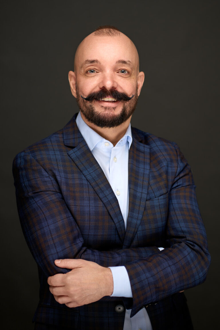 Smilende skaldet mand med stylet overskæg, iført blåternet habitjakke og lyseblå skjorte, står med armene over kors på en mørk baggrund. Perfekt til en Linkedin-portrætvirksomhed af Weigang Fotografi.