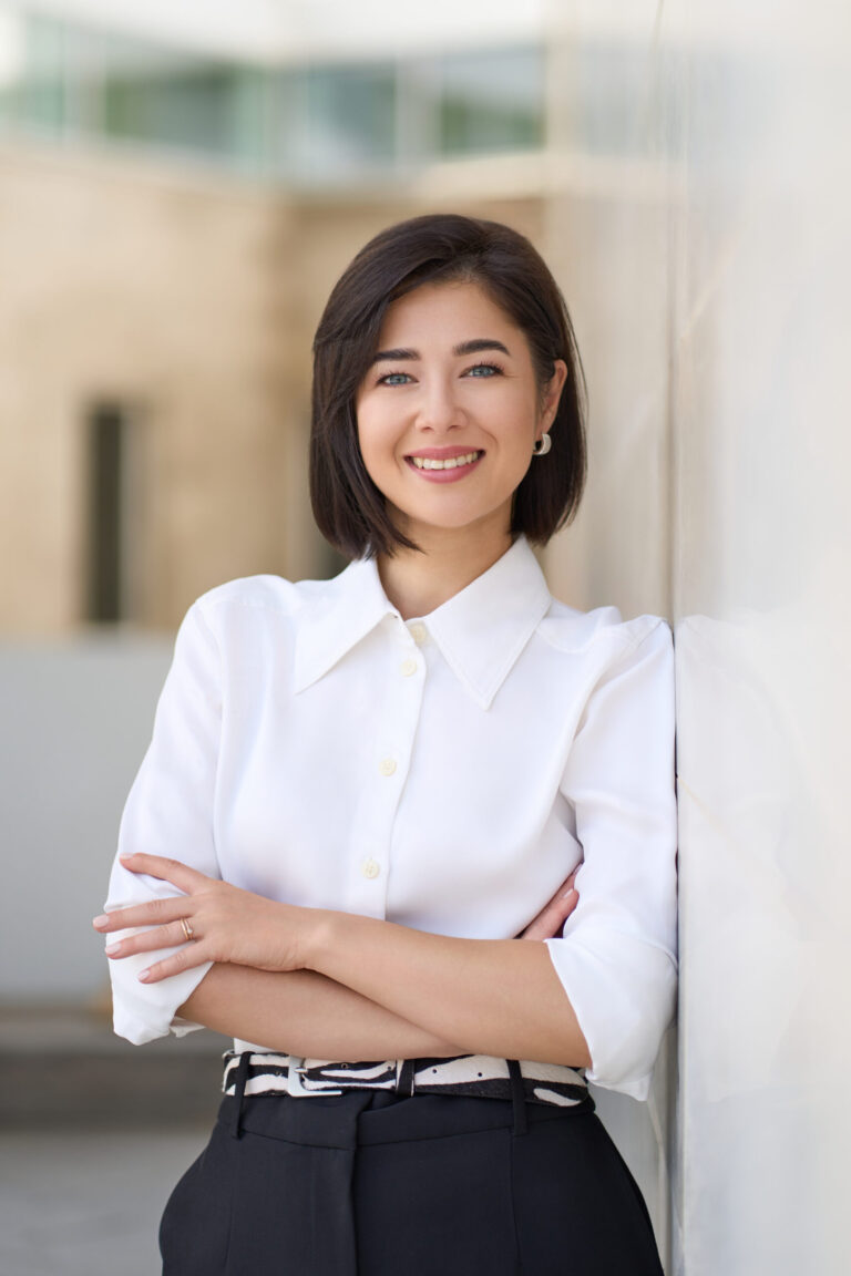 En kvinde med kort mørkt hår og en hvid bluse står udendørs, smiler selvsikkert med armene over kors og læner sig op ad en lys væg - et perfekt LinkedIn-portræt for den moderne forretningsdrivende.