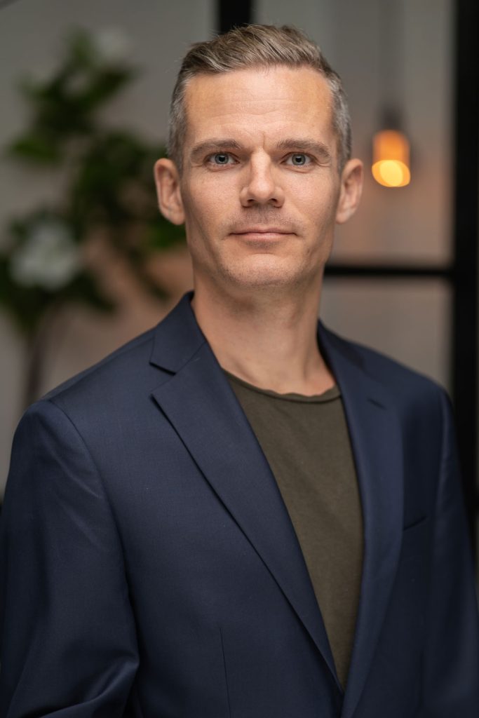 Fotograf René Beck i mørkeblå blazer og olivengrøn t-shirt i halvkropportræt med plante og varm lampe i baggrunden