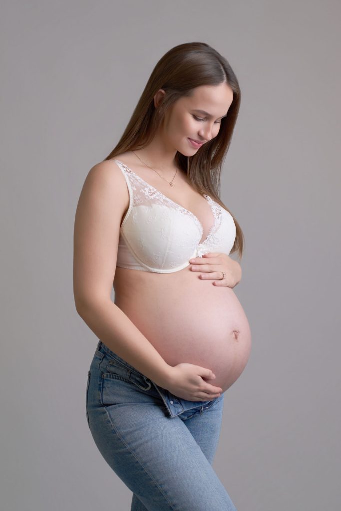 Gravid kvinde i hvid blondebh og åbne jeans holder om maven og kigger ned smilende mod en grå baggrund