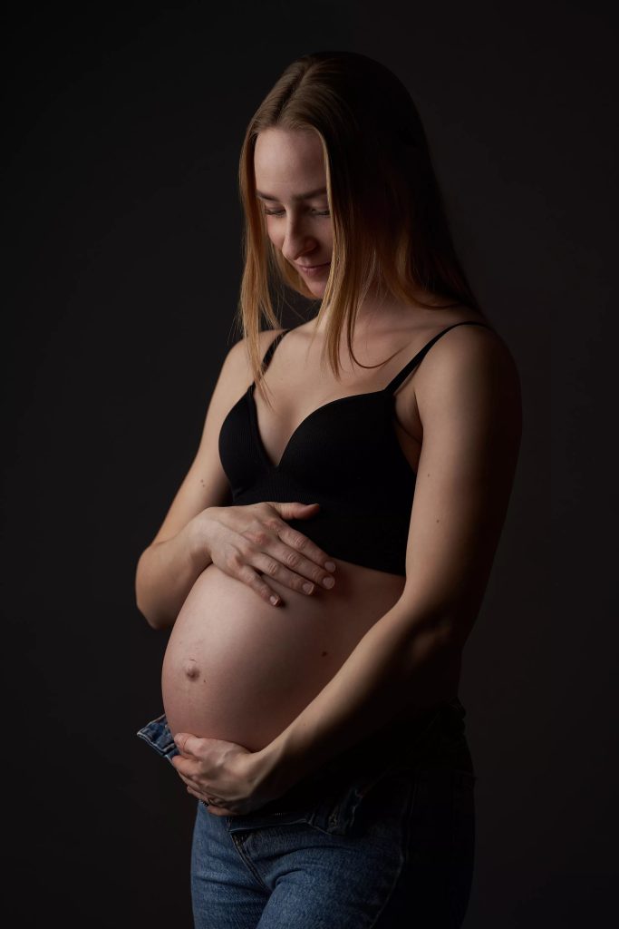 Gravid kvinde i sort bh og åbne jeans holder om sin mave, intimt studieportræt