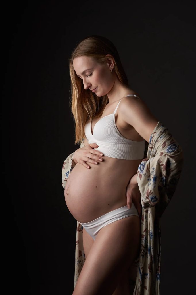 Gravid kvinde i hvidt undertøj med blomstret kimono, blødt sidelys mod sort baggrund