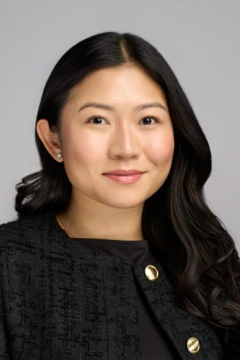 Medarbejderportræt af en professionel kvinde med langt mørkt hår, iført en sort jakke med gulddetaljer, taget i et studie med en lys grå baggrund.