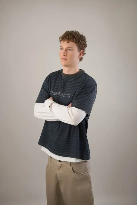 Ung mand med krøllet hår står med korslagte arme og ser lidt til siden. Han er iført et mørkeblåt stribet oversized t-shirt ovenpå en hvid langærmet trøje samt beige bukser. Studiefoto mod grå baggrund.