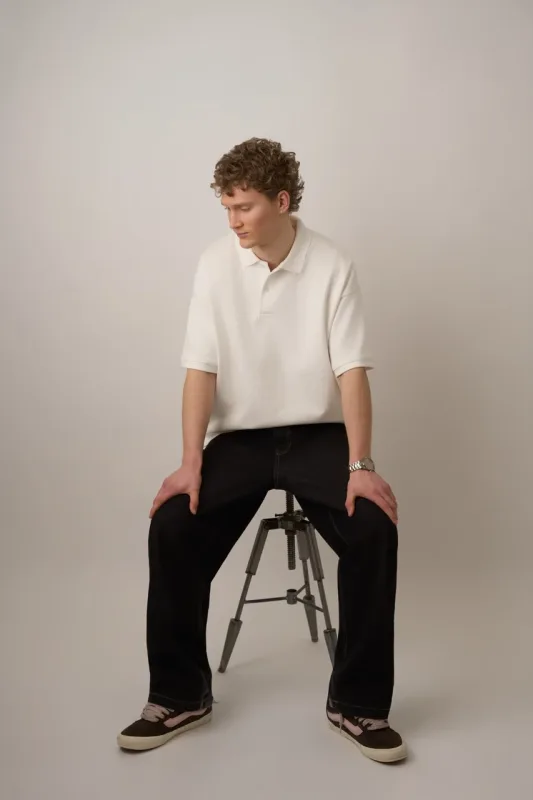 Portræt af ung mand sidder afslappet på en fotostol med hænderne på knæene og blikket rettet nedad. Iført hvid polo og brede sorte jeans med Vans sneakers og sølvur. Helkropsbillede mod lys grå baggrund.