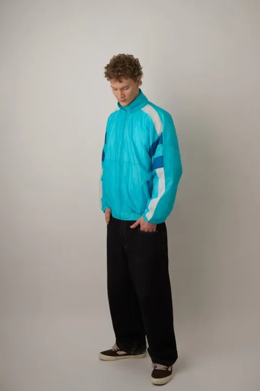 Ung mand i fuld længde iført en turkis/cyan retro windbreaker jakke med hvide og blå detaljer samt brede sorte jeans og mørke sneakers. Han kigger ned med hænderne i lommerne. Studiefoto mod grå baggrund