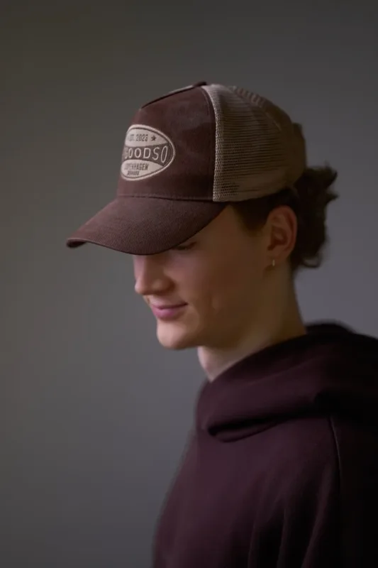Nærbillede i profil af ung kvinde med brun Goodso Copenhagen trucker cap og mørk bordeaux hoodie. Hun kigger ned med et let smil. Samme dramatiske studiebelysning som det foregående billede fremhæver kasketmærket.