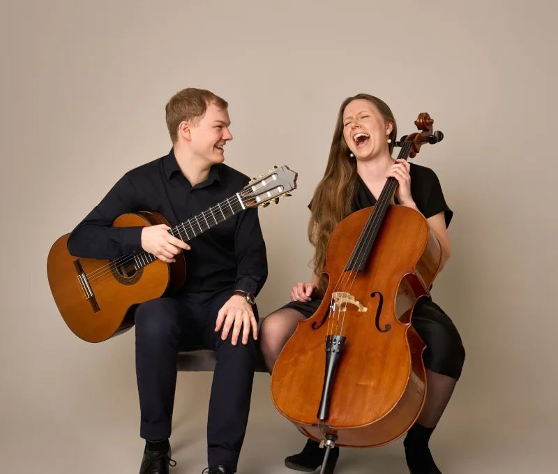 Jonas og Monica sidder side om side og griner begge højt. Han holder en klassisk guitar, hun holder en cello. Begge iført sort tøj mod varm beige baggrund.