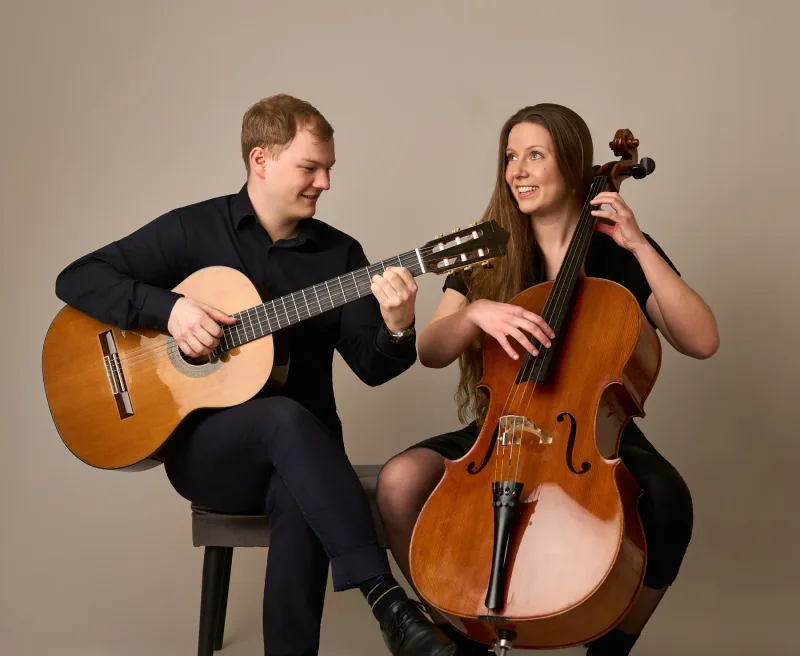 Jonas spiller klassisk guitar mens Monica holder om sin cello — begge smiler og kigger på hinanden. Naturligt og levende duoportræt mod beige baggrund.