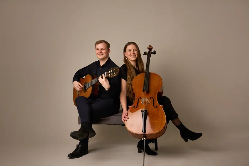 onas og Monica smiler bredt mod kameraet mens de holder deres instrumenter. Varmt og imødekommende helkropsportræt mod beige baggrund.