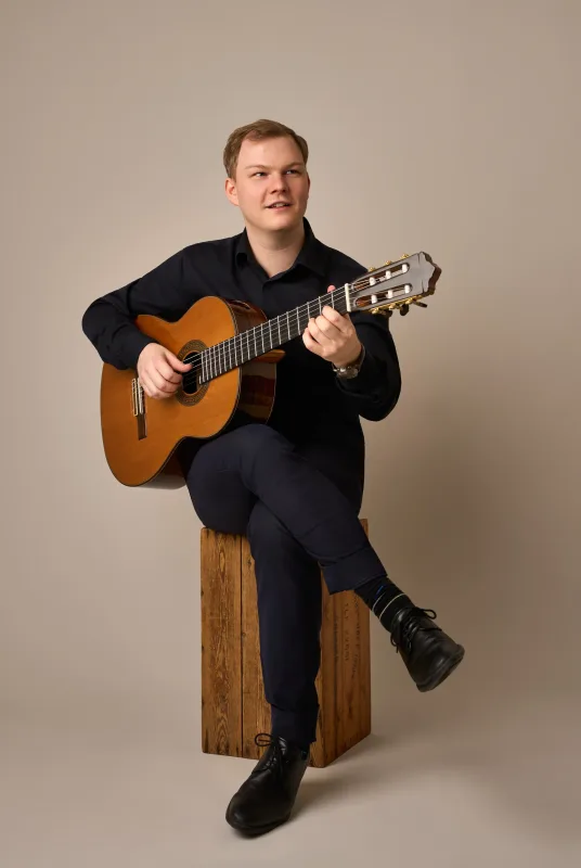 Jonas sidder på en træklods og spiller klassisk guitar med et let smil og blikket rettet opad. Helkropsportræt i sort tøj mod beige baggrund.