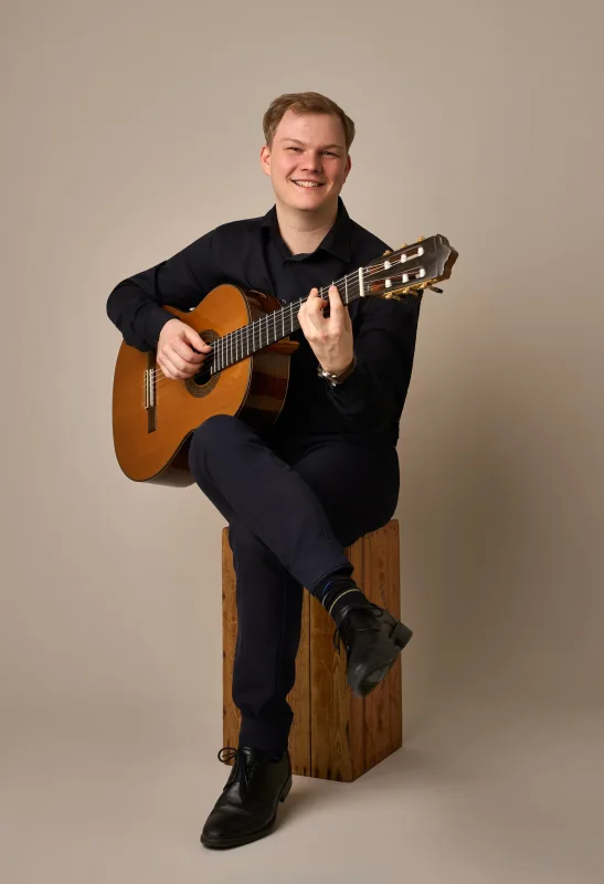 Jonas sidder på en træklods og smiler bredt mod kameraet mens han holder sin klassiske guitar. Imødekommende og energisk musikerportræt.