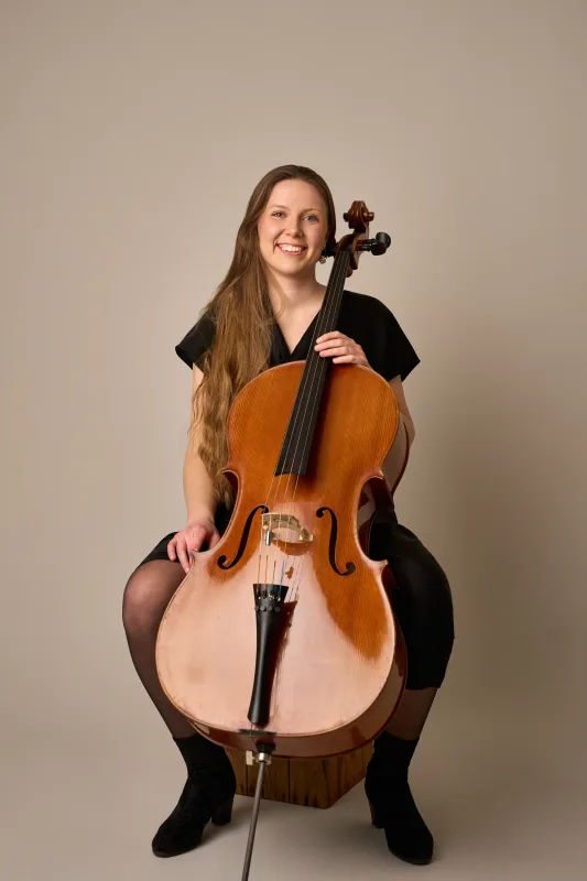 Monica sidder og smiler bredt mod kameraet mens hun holder sin cello foran sig. Varmt og venligt musikerportræt mod beige baggrund.