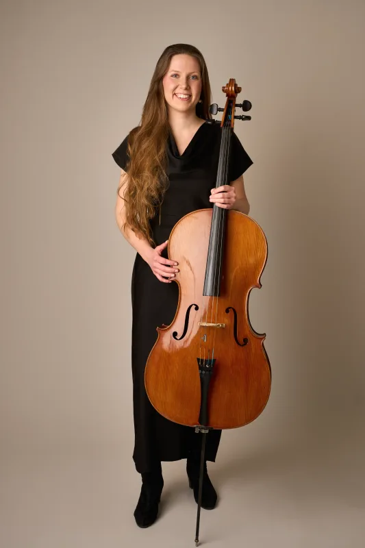 Monica står i helkrop iført sort kjole og holder sin cello ved siden af sig. Smilende og professionelt musikerportræt mod beige baggrund.