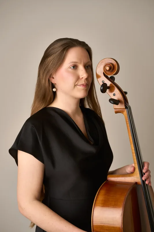 Nærbillede af Monica i halvprofil iført sort kjole med perleøreringe. Hun holder om celloen ved siden af sig og kigger roligt frem. Elegant og stilfuldt musikerportræt.