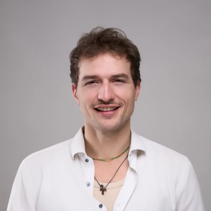 Portrætfoto af ung mand med krøllet mørkt hår, lille overskæg og åbent smil. Iført hvid skjorte med halskæde og perlehalskæde. Grå baggrund.