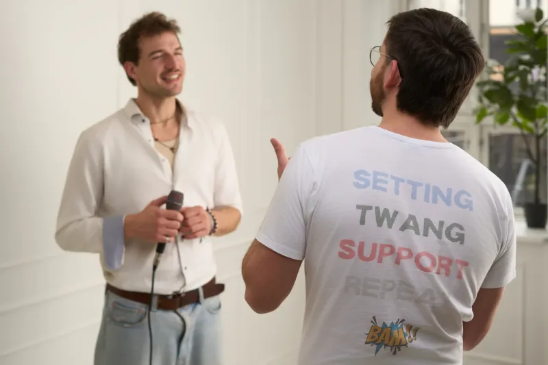 To mænd i samtale — den ene holder en mikrofon og smiler, den anden ses bagfra med en t-shirt med teksten "Setting Twang Support Repeat". Lyst interiør med store vinduer.