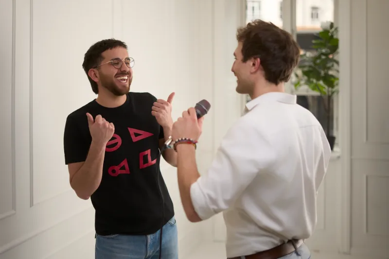 Manden med briller og sort t-shirt griner højt mens han gestikulerer. Den anden holder mikrofonen frem mod ham. Lyst lokale med vindue i baggrunden.