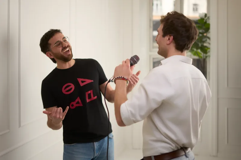 Manden med briller og sort t-shirt ler med lukkede øjne mens mikrofonen holdes frem mod ham. Afslappet og spontan stemning.